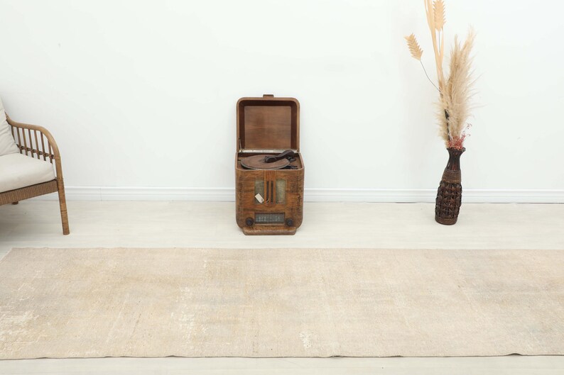 K&ouml;nnte beinhalten: Ein beigefarbener rechteckiger Teppich mit einem Used-Look. Ein offenes, vintage Holzradio steht aufrecht in der Mitte, mit einem Korbstuhl links. Eine dekorative Vase mit getrockneten Pflanzen befindet sich rechts.