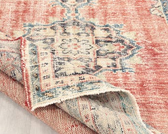 May include: A distressed, vintage-style rug in faded red and cream. The rug has a detailed, geometric pattern with blue and beige accents. The corner is folded over, revealing the underside. The rug's dimensions are in centimetres.