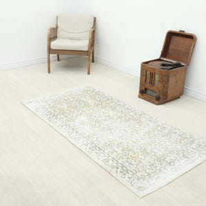 Op de afbeelding: Een rechthoekig vloerkleed met een vervaagd, verweerd patroon in tinten crème, beige en gedempt saliegroen. Het kleed heeft een gedetailleerde rand met ingewikkelde bloemmotieven en geometrische patronen, liggend op een lichte vloer in een kamer.
