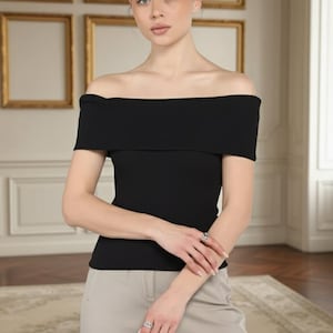 May include: Black off-the-shoulder top with short sleeves. The top has a ribbed texture and is paired with light grey trousers. The model is wearing rings on their fingers. The background features framed artwork and a patterned rug.