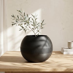May include: A black, spherical vase with vertical cut-out lines around the middle, holding a sprig of olive branches. The vase sits on a light-colored wooden table. The olive branches have green leaves and small olives.
