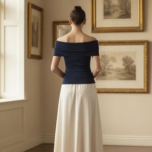 May include: A woman wearing a navy blue off-the-shoulder top and a long, cream-colored skirt. The top has short sleeves and a folded neckline. The skirt is floor-length. The woman is standing in a room with framed artwork on the wall.