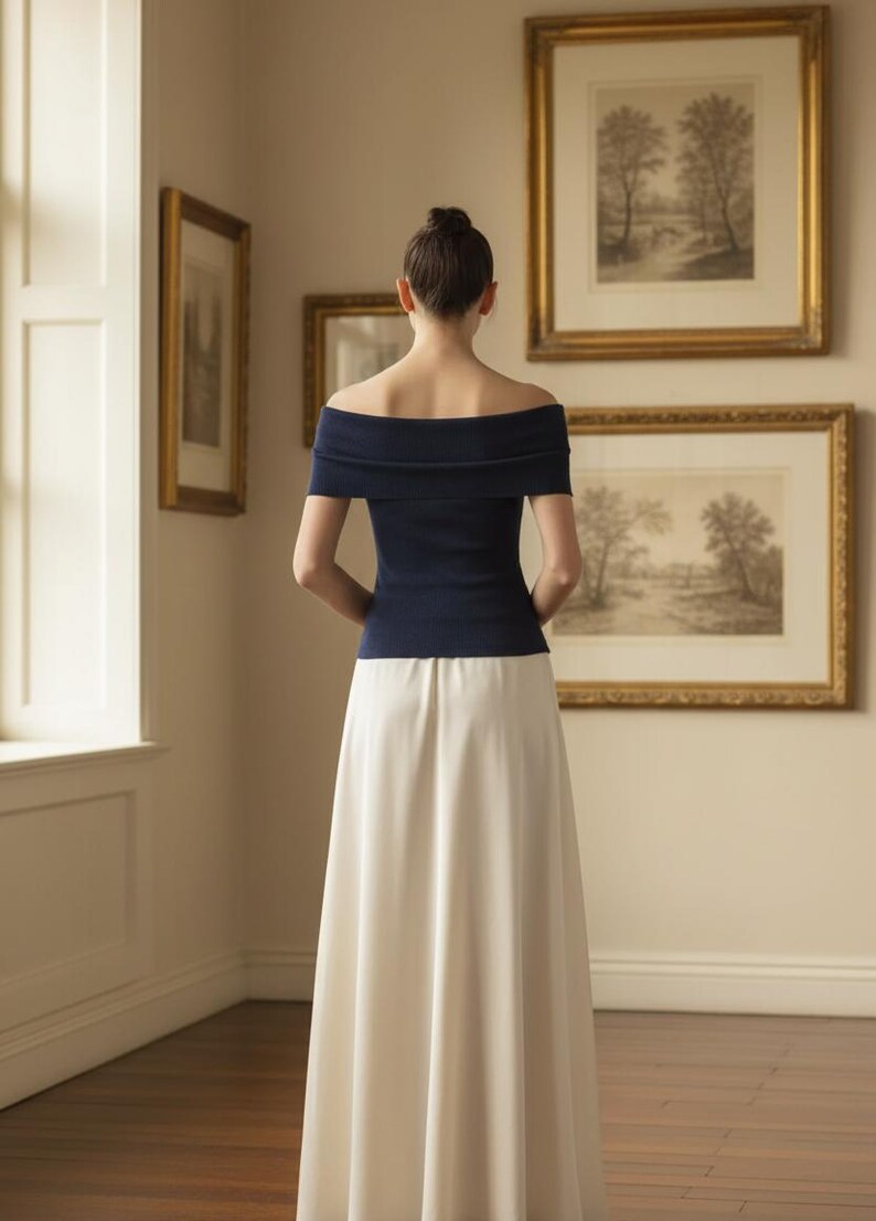 May include: A woman wearing a navy blue off-the-shoulder top and a long, cream-colored skirt. The top has short sleeves and a folded neckline. The skirt is floor-length. The woman is standing in a room with framed artwork on the wall.