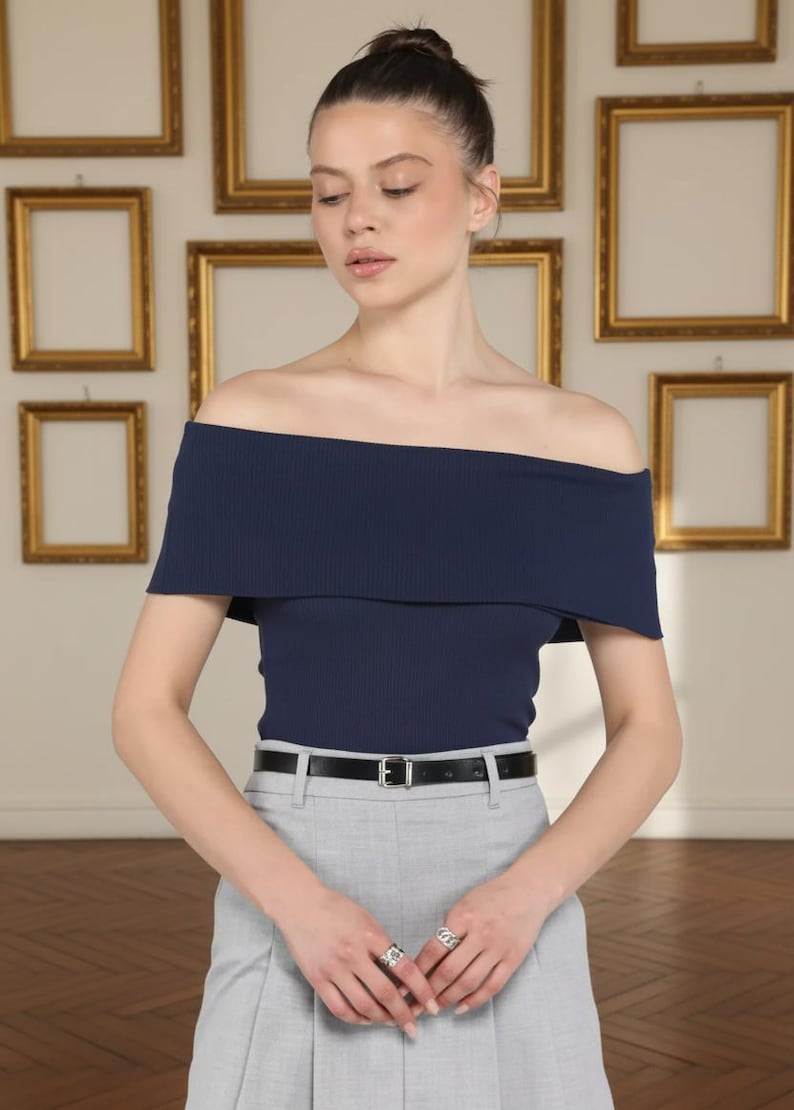 May include: A navy blue off-the-shoulder top with a ribbed texture. The top is paired with light gray trousers and a black belt. The model is wearing silver rings. The background features gold framed artwork.