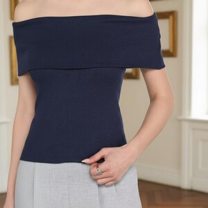 May include: A navy blue off-the-shoulder top with a ribbed texture. The top has a wide, folded-over neckline and short sleeves. The model is wearing gray pants. The background features framed artwork.