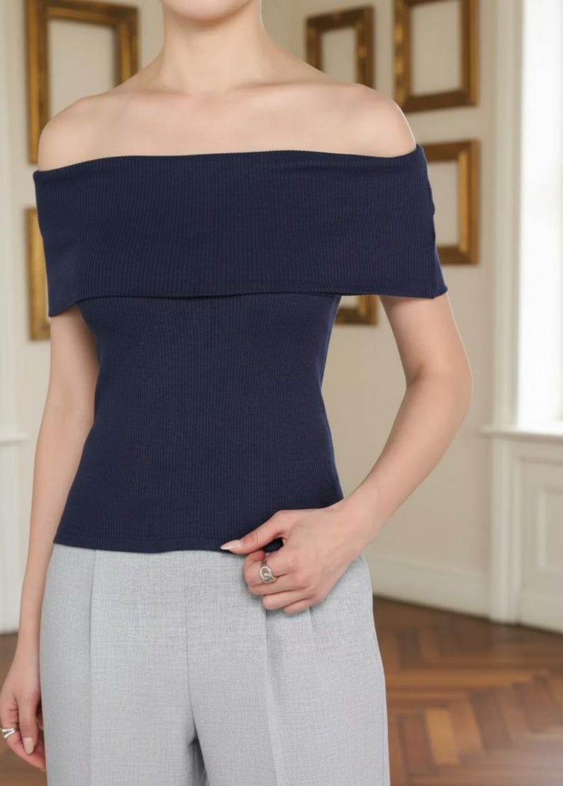 May include: A navy blue off-the-shoulder top with a ribbed texture. The top has a wide, folded-over neckline and short sleeves. The model is wearing gray pants. The background features framed artwork.