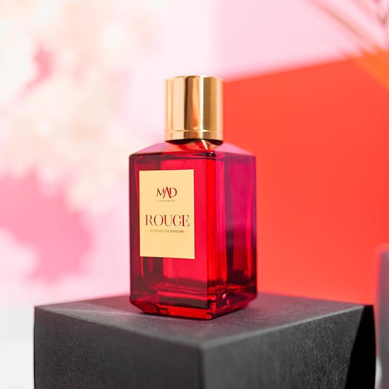 May include: A red perfume bottle with a gold cap and a rectangular shape. The bottle has a label that reads "MAD ROUGE". The perfume bottle is sitting on a black surface.