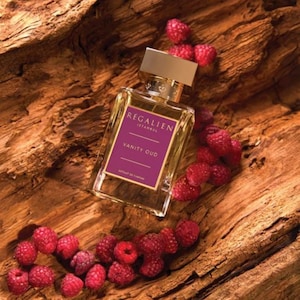 May include: A rectangular perfume bottle with a gold cap and a purple label that reads "REGALIEN ISTANBUL VANITY OUD". The bottle is surrounded by fresh red raspberries, all set against a textured wooden background.