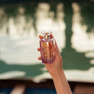 May include: A rectangular perfume bottle with a clear, faceted top and a purple base is held in a hand. The bottle has a gold label that reads "Mona Lisa" in cursive script. The bottle is filled with a light-colored liquid.