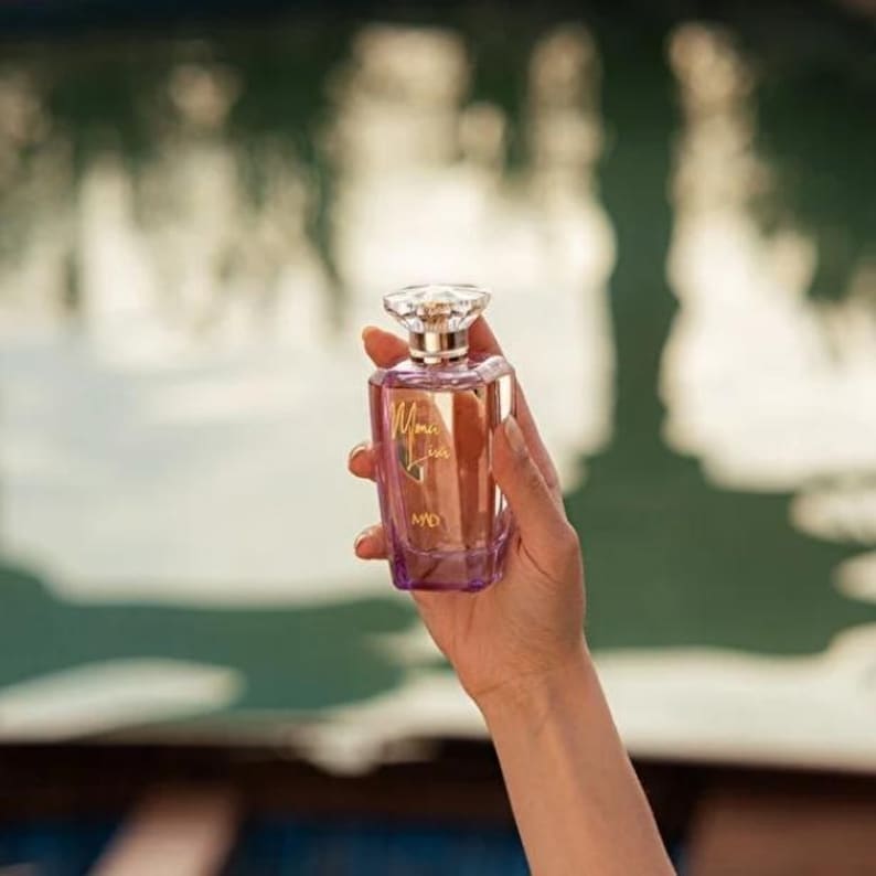 May include: A rectangular perfume bottle with a clear, faceted top and a purple base is held in a hand. The bottle has a gold label that reads "Mona Lisa" in cursive script. The bottle is filled with a light-colored liquid.