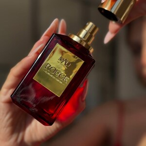 May include: A rectangular, ruby-red perfume bottle with a gold label and cap. The label reads "MAD ROUGE" in gold lettering. The bottle is held in a person's hands, with the cap being removed. The background is blurred.