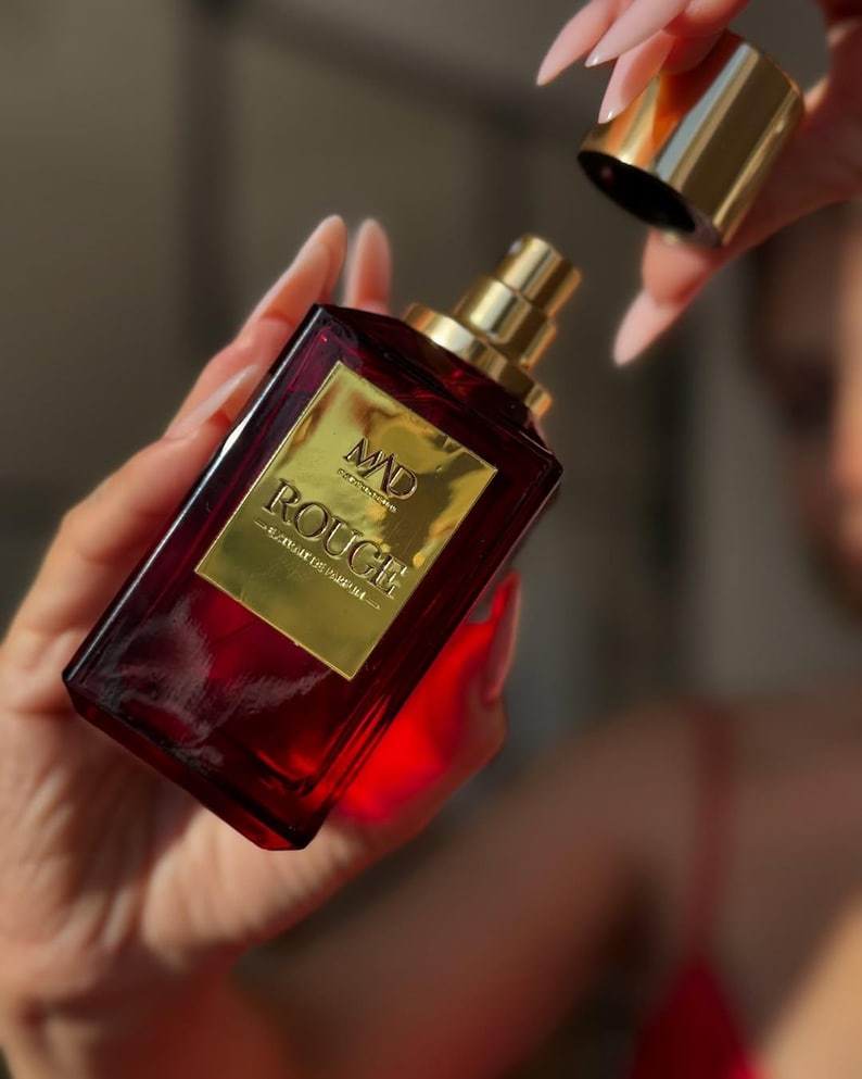 May include: A rectangular, ruby-red perfume bottle with a gold label and cap. The label reads "MAD ROUGE" in gold lettering. The bottle is held in a person's hands, with the cap being removed. The background is blurred.
