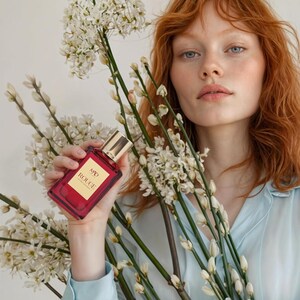 May include: A red perfume bottle with gold accents and the text "MAD ROUGE" is held in front of white flowers. The bottle is rectangular. The person in the image is wearing a light blue shirt and has red hair.