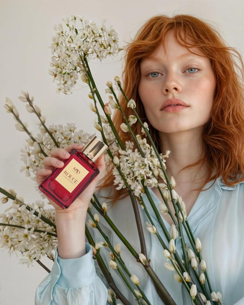 May include: A red perfume bottle with gold accents and the text "MAD ROUGE" is held in front of white flowers. The bottle is rectangular. The person in the image is wearing a light blue shirt and has red hair.