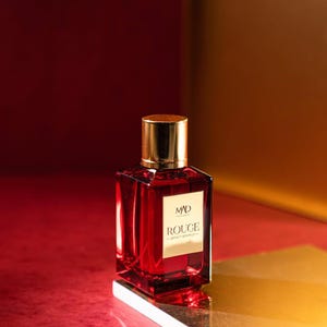 May include: A red perfume bottle with a gold cap and a gold label that reads "MAD ROUGE". The bottle is rectangular and filled with a red liquid. The background is a gradient of red and gold.