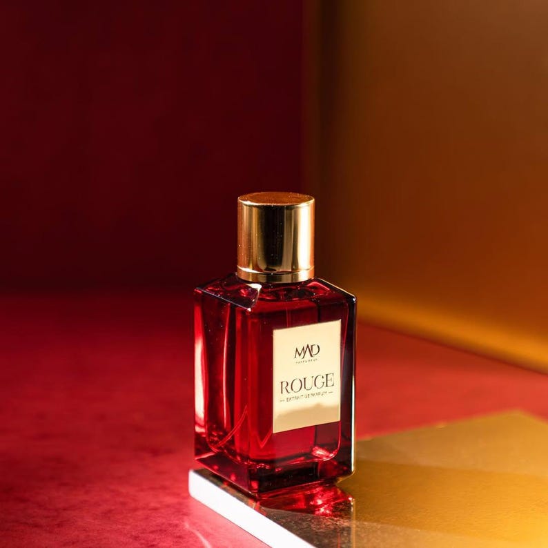 May include: A red perfume bottle with a gold cap and a gold label that reads "MAD ROUGE". The bottle is rectangular and filled with a red liquid. The background is a gradient of red and gold.