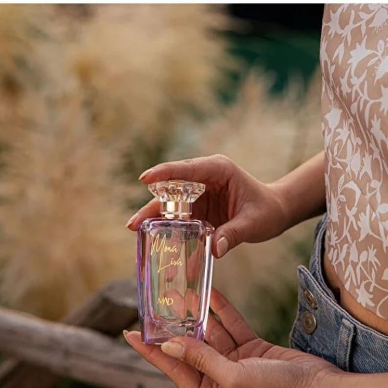 May include: A rectangular perfume bottle with a clear, iridescent body is held in two hands. The bottle has a crystal-like stopper and gold lettering that reads "Mona Lisa" and "MD". The background is blurred with neutral tones.