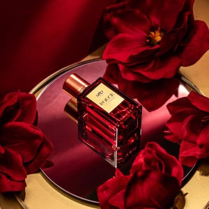 May include: A red perfume bottle with a gold cap and label, labeled "MD ROUGE", sits on a reflective surface surrounded by red roses. The bottle has a rectangular shape with a textured design, and the background is a deep red.