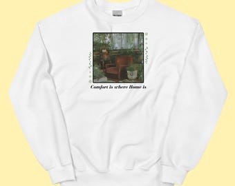 Può includere: Felpa bianca con un'immagine incorniciata di un accogliente spazio abitativo. L'immagine presenta una poltrona marrone, piante e la scritta "Comfort is where Home is". Accenti verdi ed elementi decorativi circondano l'immagine.