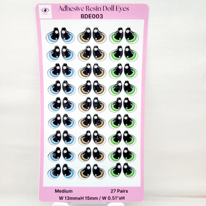 May include: A sheet of 27 pairs of adhesive resin doll eyes. The eyes are medium size and come in three colors: blue, brown, and green. The sheet is labeled "Adhesive Resin Doll Eyes BDE003" and "Medium W 13mmxH 15mm / W 0.51"xH".