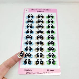 May include: A sheet of 27 pairs of medium-sized adhesive resin doll eyes. The eyes are black with white and either blue, gold, or green accents. The sheet is labeled "BDE003" and "W 13mmxH 15mm / W 0.51"xH 0.59"".