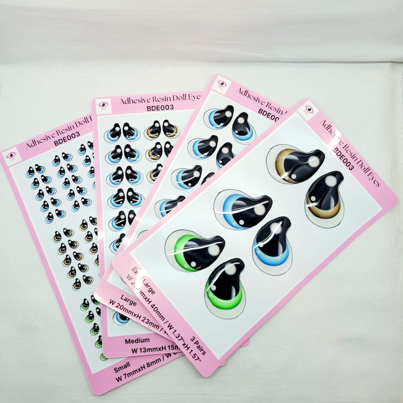 May include: Three sheets of adhesive resin doll eyes in various sizes. The sheets are pink with white borders and feature different colored eyes. The sizes are labeled as Ex Large, Large, Medium, and Small. The measurements are listed in inches and millimeters.