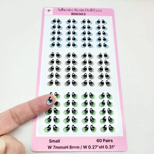 May include: A sheet of 60 pairs of small adhesive resin doll eyes. The eyes are black with a white iris and a variety of colored outer rings, including blue, beige, and green. The sheet is labeled "Adhesive Resin Doll Eyes BDE003" and "Small W 7mmxH 8mm / W 0.27"xH 0.31".