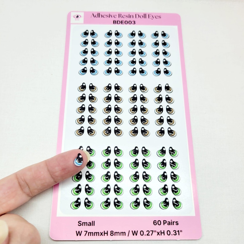 May include: A sheet of 60 pairs of small adhesive resin doll eyes. The eyes are black with a white iris and a variety of colored outer rings, including blue, beige, and green. The sheet is labeled "Adhesive Resin Doll Eyes BDE003" and "Small W 7mmxH 8mm / W 0.27"xH 0.31".