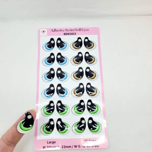 May include: A sheet of 12 pairs of large adhesive resin doll eyes. The eyes are black with a white iris and come in blue, brown, and green colors. The sheet is labeled "Adhesive Resin Doll Eyes BDE003" and "Large W 20mm 23mm/W 0.70 . 0.50".