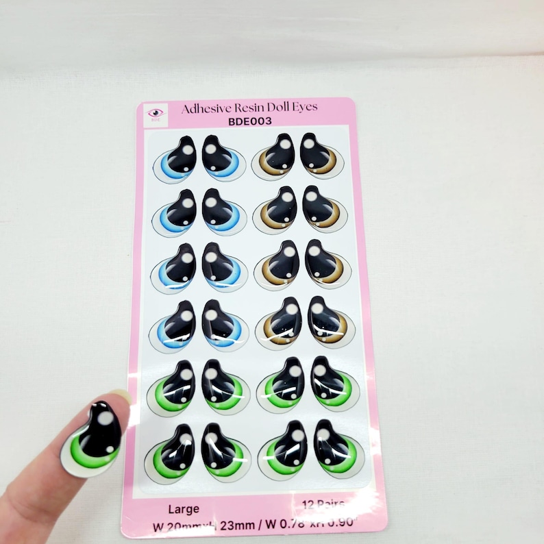 May include: A sheet of 12 pairs of large adhesive resin doll eyes. The eyes are black with a white iris and come in blue, brown, and green colors. The sheet is labeled "Adhesive Resin Doll Eyes BDE003" and "Large W 20mm 23mm/W 0.70 . 0.50".