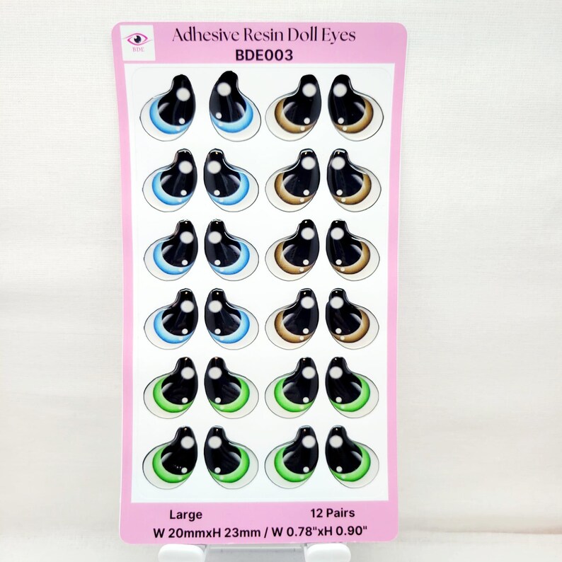 May include: A sheet of 12 pairs of large adhesive resin doll eyes. The eyes are black with a white iris and a blue, green, or brown outer ring. The sheet is pink and white and has the text "Adhesive Resin Doll Eyes BDE003 Large 12 Pairs W 20mmxH 23mm / W 0.78"xH 0.90"".