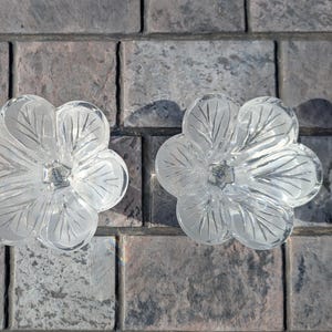 May include: Two clear glass flower-shaped candle holders. Each has five petals with etched leaf details. The candle holders are on a gray and brown brick surface. The glass is transparent, allowing light to pass through.