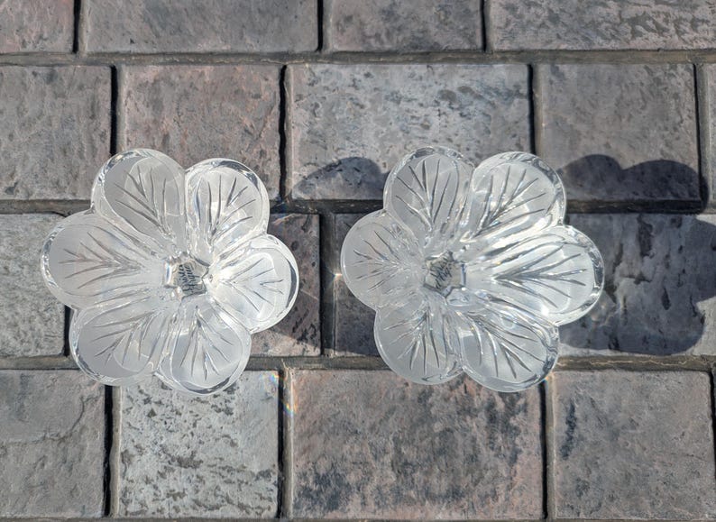 May include: Two clear glass flower-shaped candle holders. Each has five petals with etched leaf details. The candle holders are on a gray and brown brick surface. The glass is transparent, allowing light to pass through.