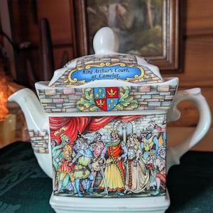 May include: A white ceramic teapot with a brick-like design and a scene of King Arthur's court at Camelot. The teapot has a lid with a gold crown and the words "King Arthur's Court, at Camelot."