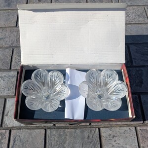 May include: Two clear, flower-shaped glass candle holders inside a cardboard box. The candle holders have a frosted leaf design and are resting on a dark blue surface. The box is open, revealing the contents.