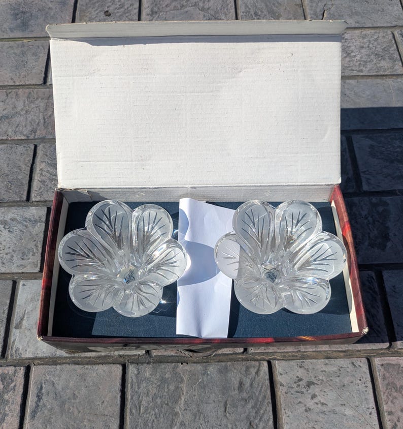 May include: Two clear, flower-shaped glass candle holders inside a cardboard box. The candle holders have a frosted leaf design and are resting on a dark blue surface. The box is open, revealing the contents.