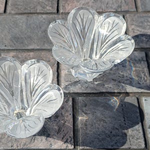 May include: Two clear glass flower-shaped candle holders. Each holder has a detailed petal design with etched lines. The candle holders are sitting on a gray brick surface, with shadows cast by the sunlight.