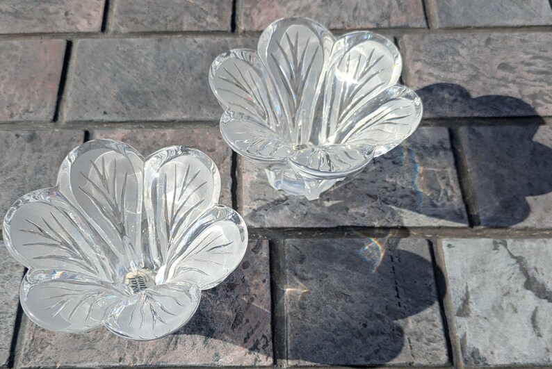 May include: Two clear glass flower-shaped candle holders. Each holder has a detailed petal design with etched lines. The candle holders are sitting on a gray brick surface, with shadows cast by the sunlight.