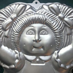May include: A silver metal cake pan shaped like a girl with pigtails and a bow. The pan has a detailed face with eyes, nose, and mouth.