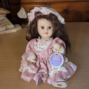 May include: A porcelain doll with brown hair and a pink dress. The doll has a pearl necklace and a tag that reads "Year 2002 Edition by Christina Verdi". The dress has white lace trim and the doll is wearing a matching bonnet.