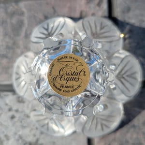 May include: Clear crystal glass with a floral design. The glass has a gold sticker that reads "Cristal d'Arques France Genuine Lead Crystal". The glass has a flower-like shape with six petals.