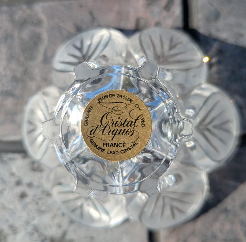 May include: Clear crystal glass with a floral design. The glass has a gold sticker that reads "Cristal d'Arques France Genuine Lead Crystal". The glass has a flower-like shape with six petals.