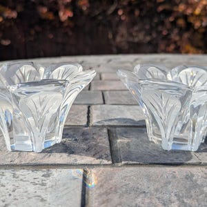 May include: Two clear glass candle holders with a floral design. Each holder has a petal-like shape with etched leaf patterns. The candle holders are on a gray stone surface, with a blurred background of foliage.