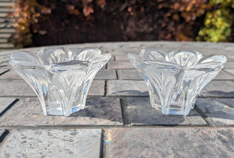 May include: Two clear glass candle holders with a floral design. Each holder has a petal-like shape with etched leaf patterns. The candle holders are on a gray stone surface, with a blurred background of foliage.