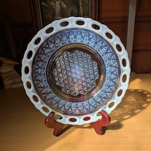 May include: A vintage, iridescent glass plate with a white, openwork rim and a geometric pattern in the center. The plate is displayed on a wooden stand, showcasing its intricate design and color variations.