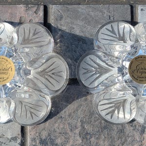 May include: Two clear, flower-shaped glass coasters with etched leaf designs. Each coaster has a gold label in the center with the text "Cristal d'Arques France". The coasters are on a gray stone surface.