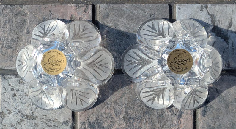 May include: Two clear, flower-shaped glass coasters with etched leaf designs. Each coaster has a gold label in the center with the text "Cristal d'Arques France". The coasters are on a gray stone surface.