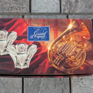 May include: A box featuring two clear, flower-shaped candle holders. The box has a red and brown background with a golden French horn illustration and the text "Cristal d'Arques" in a blue square.