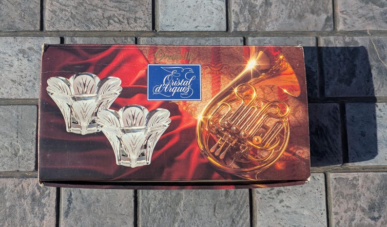 May include: A box featuring two clear, flower-shaped candle holders. The box has a red and brown background with a golden French horn illustration and the text "Cristal d'Arques" in a blue square.