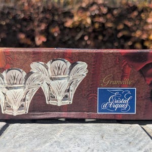 May include: A rectangular box with a maroon and red design featuring two clear crystal candle holders. The box has the text "Granville" and a blue label with "Cristal d'Arques". A gold seal reads "24% PBO".